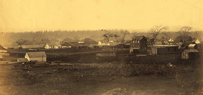 Sepia toned photograph of a set of wooden framed buildings with trees in the background  and grassland in the foreground.