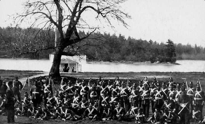 black and white photograph of a large number of men in military uniform gathered in front of a tree in front of a bay.