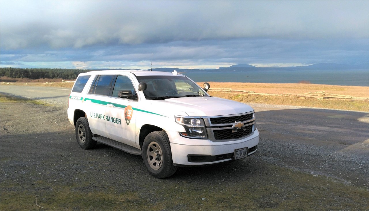 Color photograph of an SUV with the words National Park Service on the side in front of dramatically colored skies.