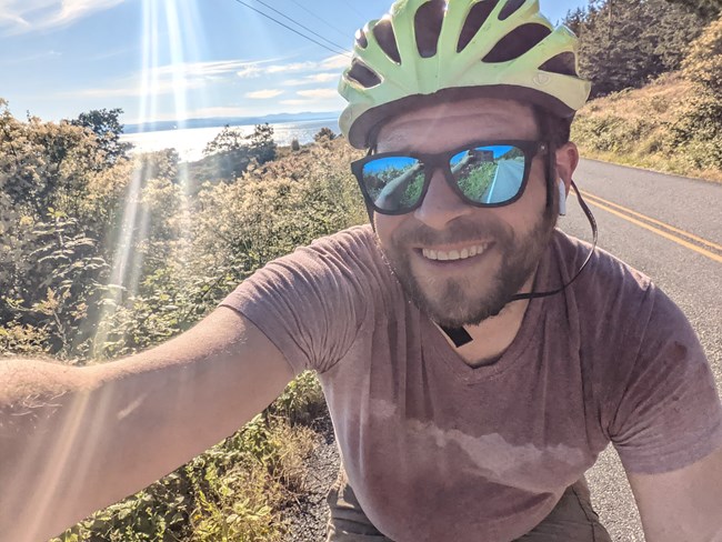 A man on a bike takes a selfie on a road. He's wearing reflective sunglasses and a lime green bike helmet. Behind him is green land, and a sliver of water. It is very sunny