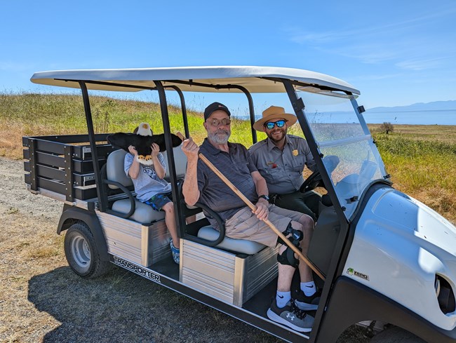 A ranger is riding a golf cart with two visitors. The visitor in the front has a walking stick and knee brace