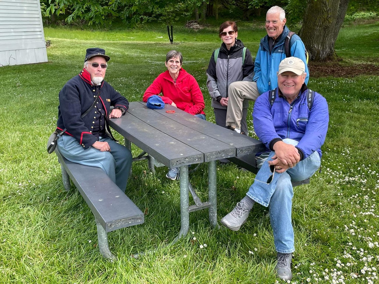 five people sit and stand around a picnic table with benches. one of them is dressed in a historical military uniform