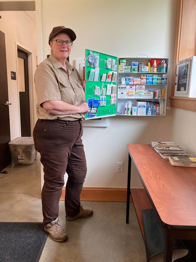 a feminine presenting park volunteer points to a fully stocked first aid box that is hanging on the wall