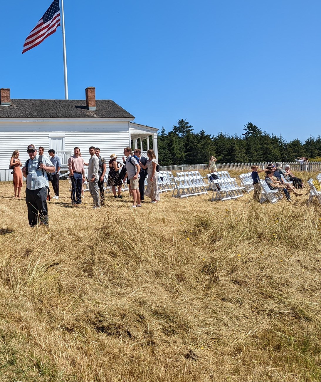 A group of well dressed visitors sitting in white chairs on a prairie in front of a whitewashed historic wood frame building with a giant american flag behind it.