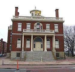 The Custom House is a large, square two story building with a cupola in the middle of the roof and a large portico over the grand stairs that sweep up to the front door.