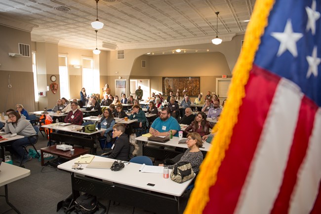 Large room full of people sitting at tables and facing forward. Stars and stripes from the American flag out of focus in foreground. Large room full of people sitting at tables and facing forward. Stars and stripes from the American flag out of focus in foreground.