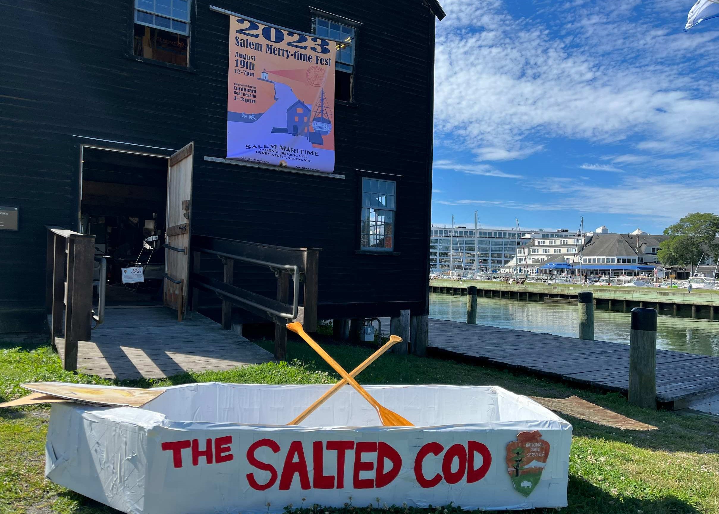 SaltedCod2023 eight foot white cardboard boat with two oars in front of a brown building next to the water