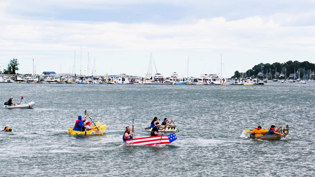 Four teams of two paddle homemade cardboard boats in a harbor