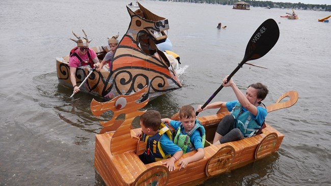 Two teams in homemade cardboard boats paddle in a harbor