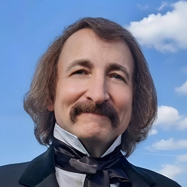 Portrait of a man with shoulder-length hair and a mustache, dressed in a black suit and bow tie against a blue sky.