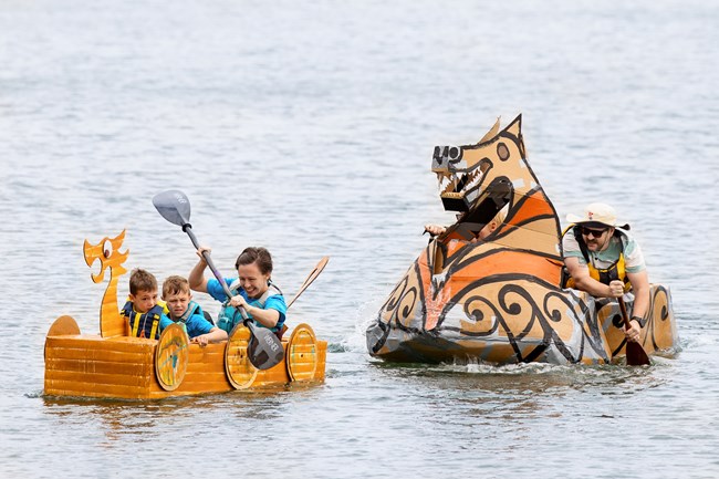 Two teams in homemade cardboard boats paddle in a harbor