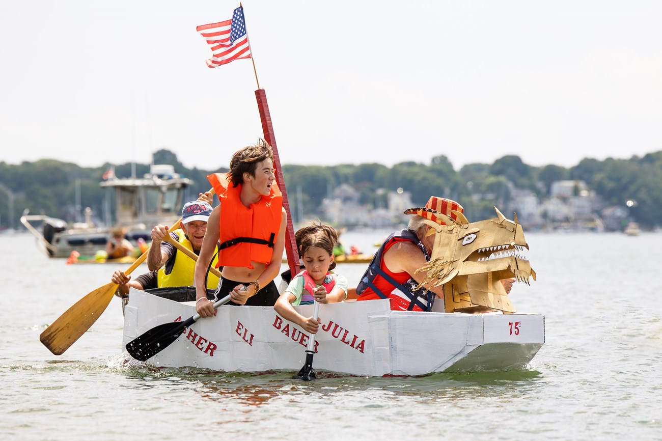 A family paddles a boat made of cardboard for a fun race in the harbor.
