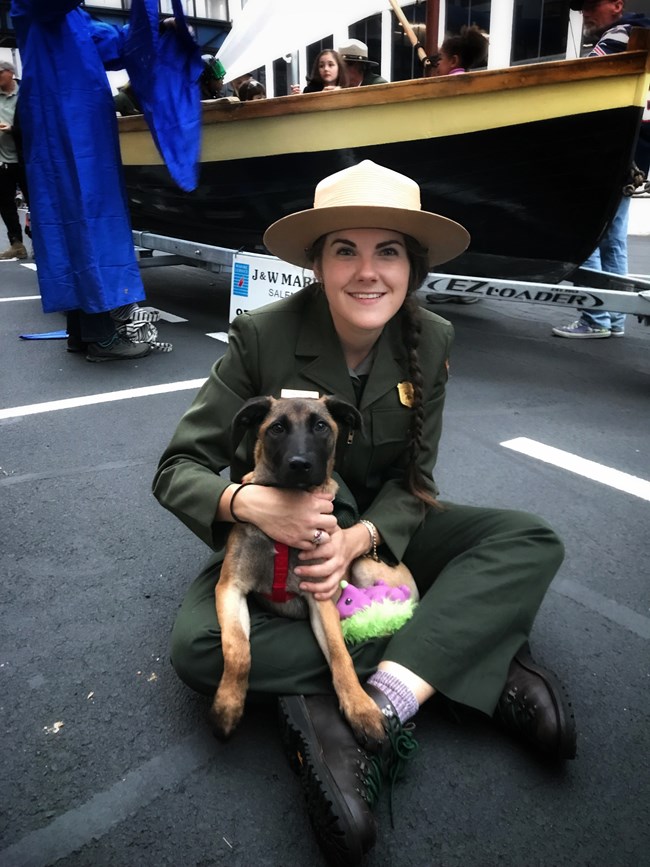 park ranger in green pants and jacket with tan flat hat sits on ground holding a dog in their lap