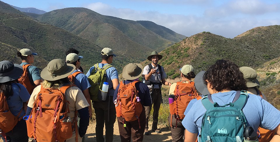 Group of youth gathered around a ranger in front of mountains.