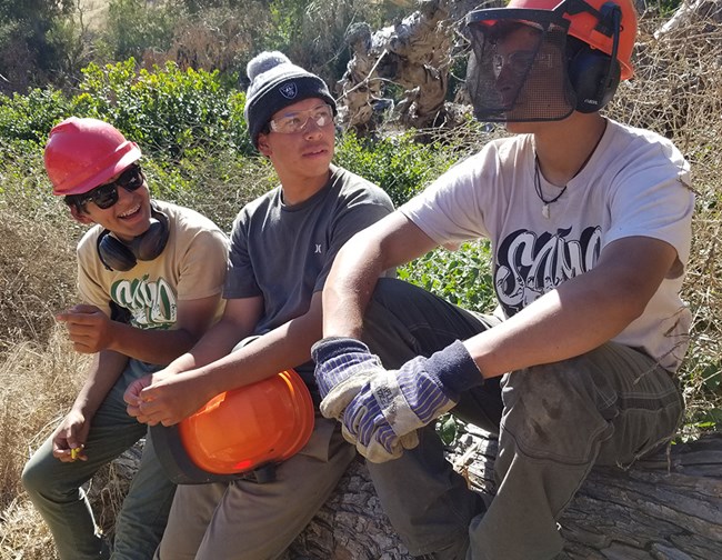 Three youth sitting on a log with hard hats and work gloves laughing.