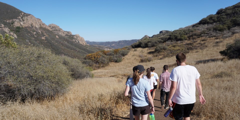 Group of hikers on the right follow a dirt path through vegetation with the mountains in the background
