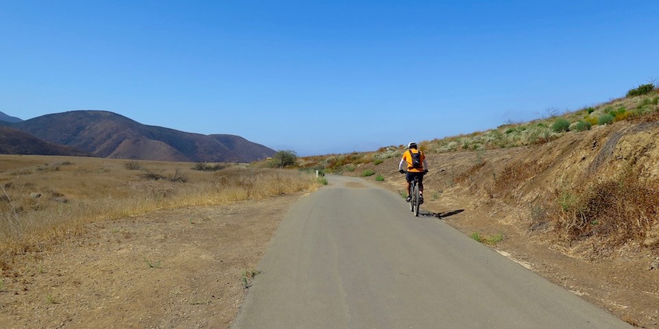 Biker to the right on small road surrounded by grassland with mountains in the background