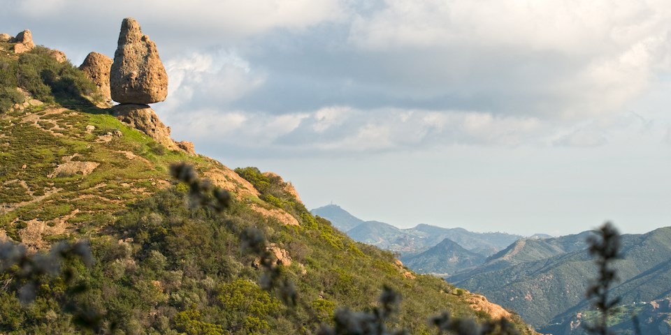 A large rock balances precariously on a cliff while mountain ridges recede into the background