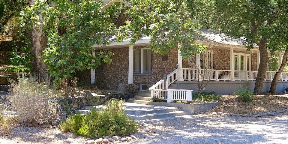 Steps lead up to a white porch and a house overlaid with stones surrounded by trees and greenery