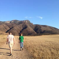Two hikers walk along a trail through an open meadow with large mountains in the background