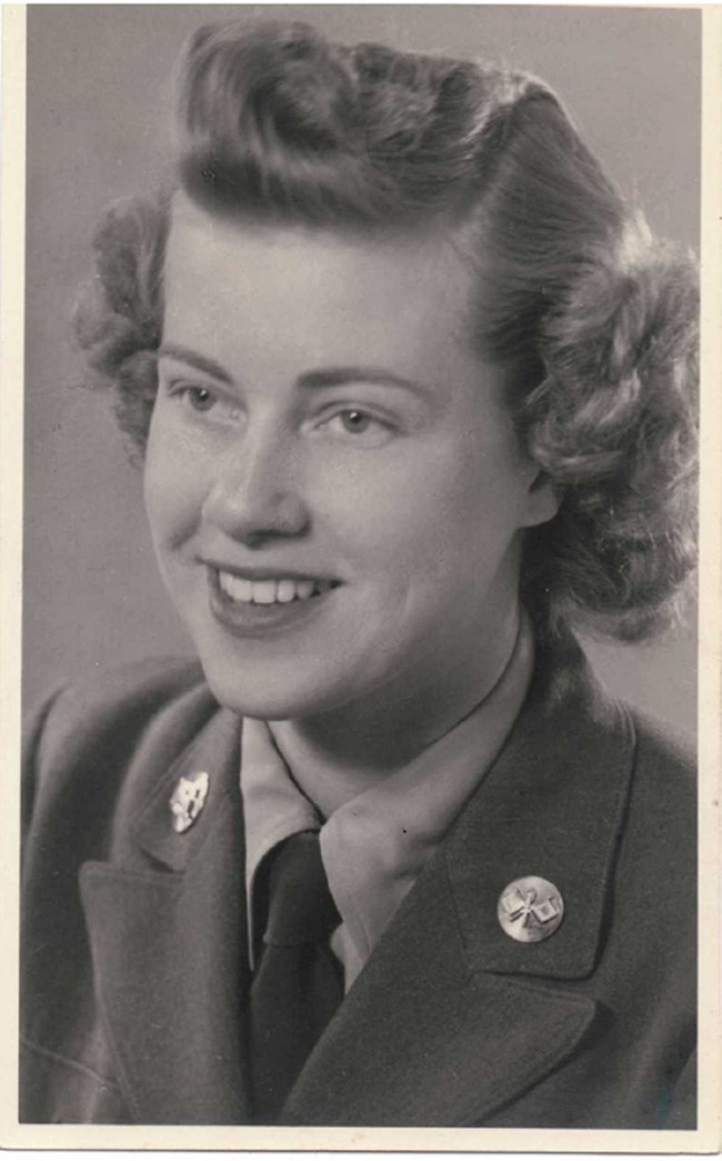 Woman in army uniform, with tie, smiling, curly hair, from neck up.