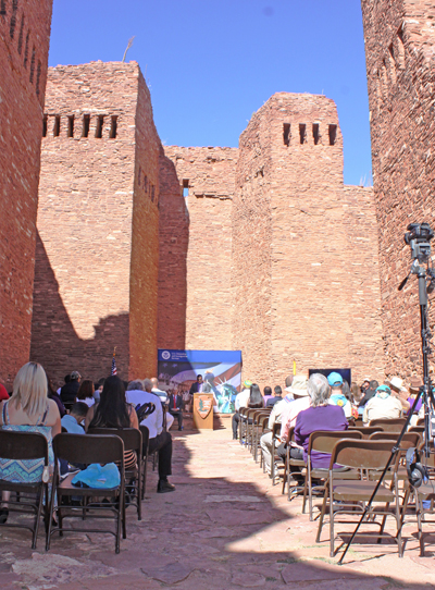 People in folding chairs sit inside the ruins of a stone mission listening to a speaker at the podium.