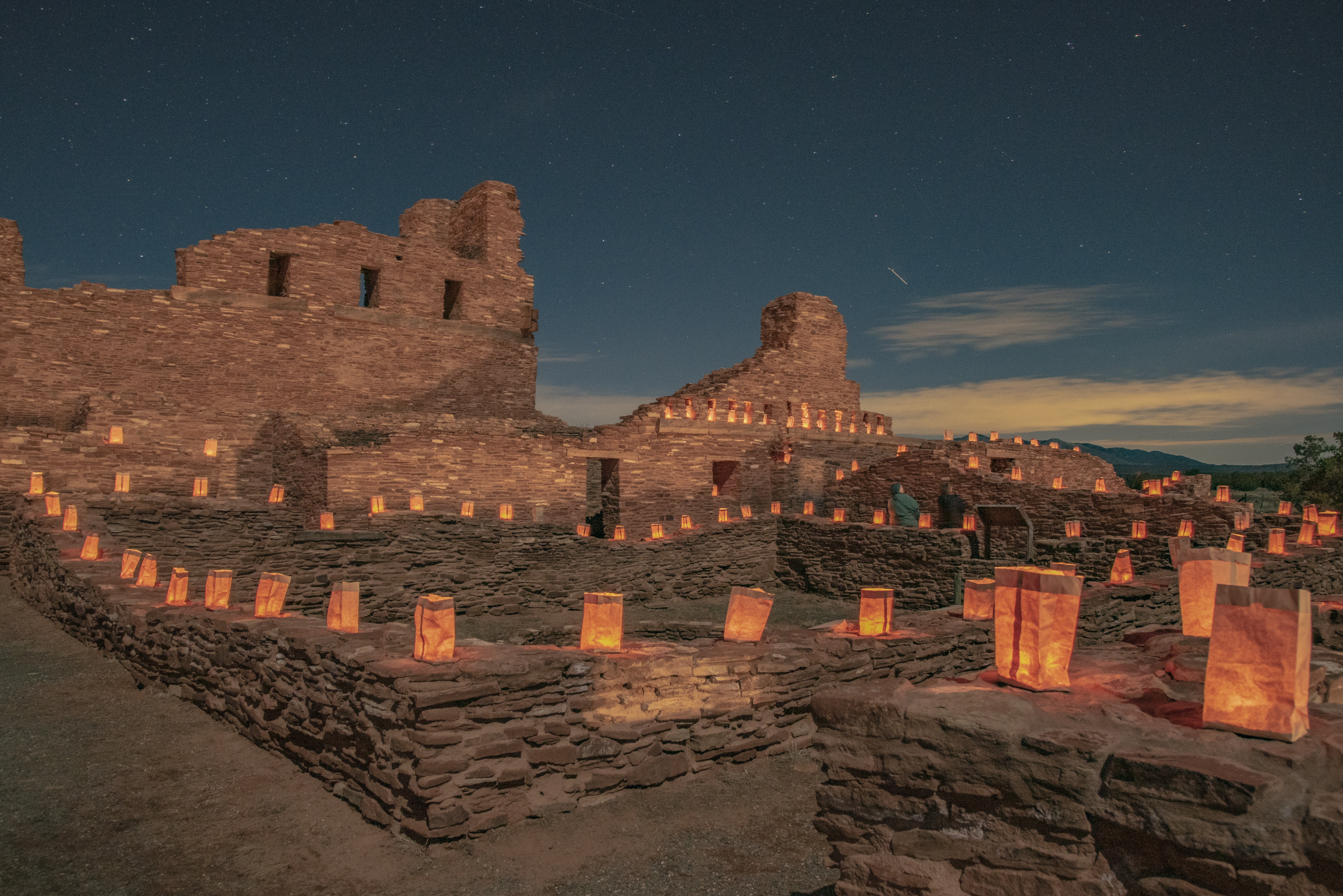Historic stone Spanish mission walls lit by glowing luminarias under a starry night sky.