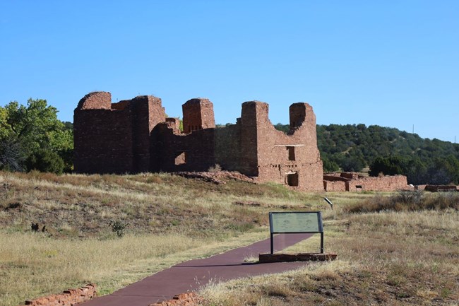 Maroon trail with wayside exhibit next to it in the center of the image. The red sandstone church at Quarai ruins in the background.