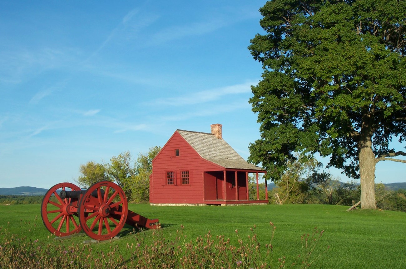 A red house sits next to a tall, green tree. It has a plush green yard surrounding it with a red canon in the foreground.