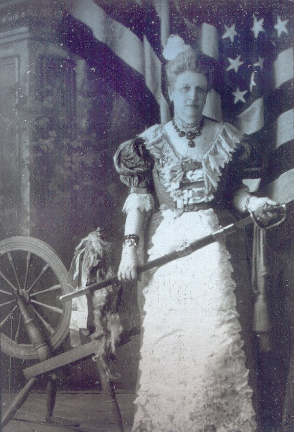 Woman in an ornate dress and holding a sheathed Revolutionary War sword.