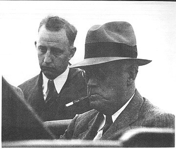 President Roosevelt sits in his car studying some papers while another man, over his right shoulder, looks on.
