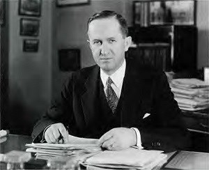 A man in a suit sits at a desk with a pen in hand and a pile of papers on the desk.