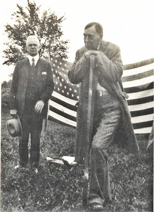 Two men in suits, one (left) watches as another (right) steps and leans on a shovel. An American flag hangs horizontally behind them