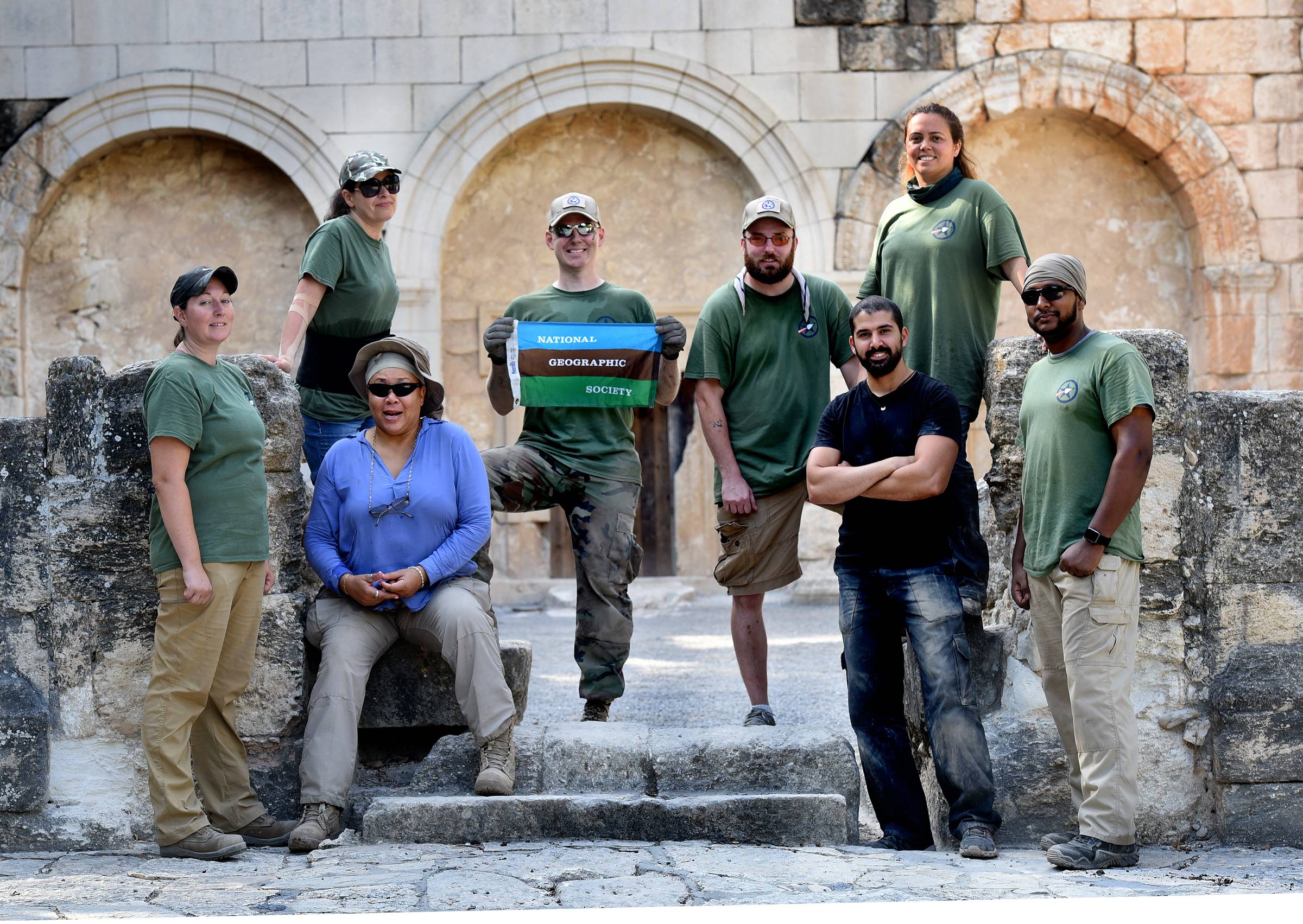 group of 8 military veterans in work clothes standing on ancient stairs and holding a small national geographic society flag