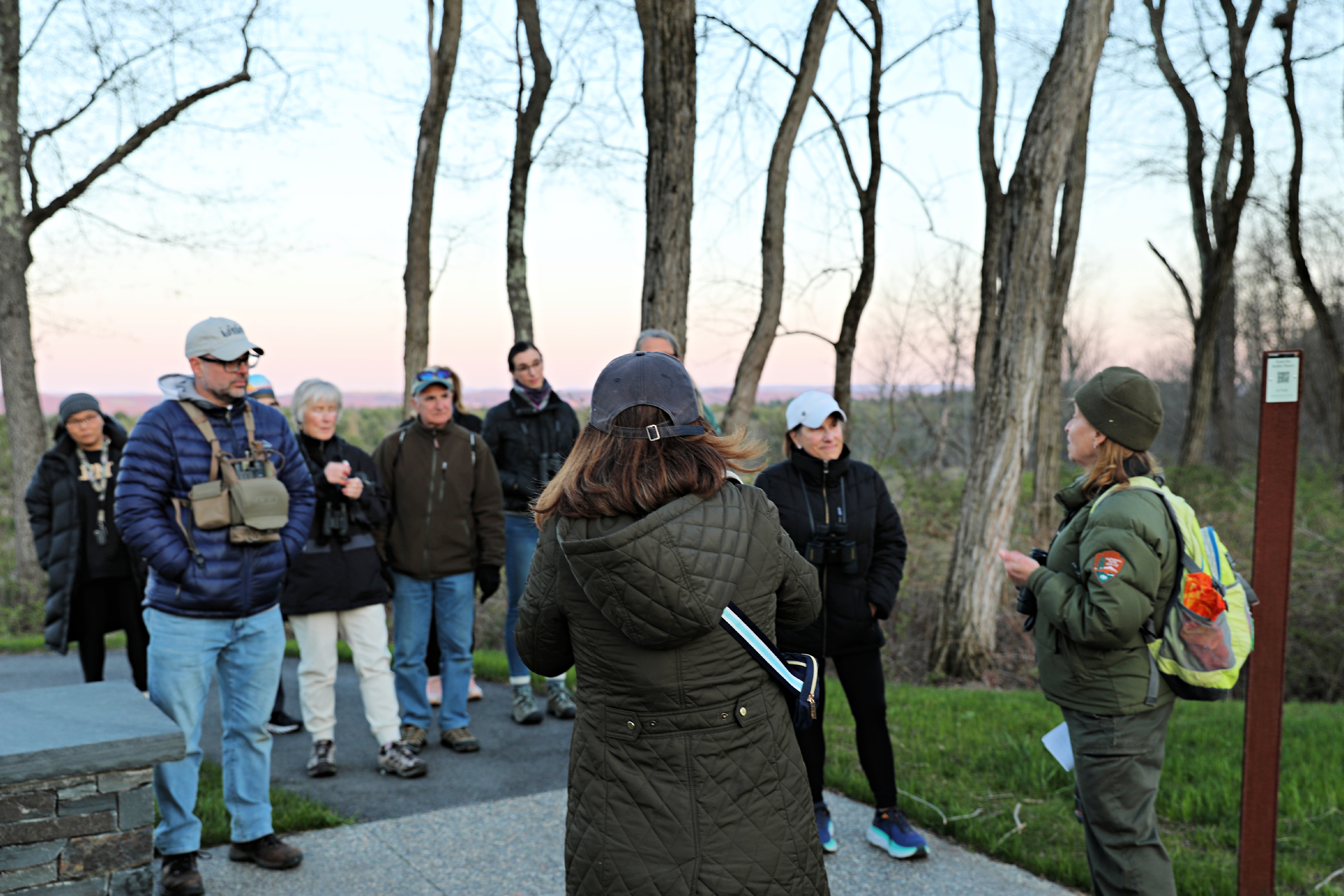 A Park Ranger leads a guided nature walk. A woman in a park ranger uniform leads a group of people on a nature hike. They stand on a hill overlooking a field.