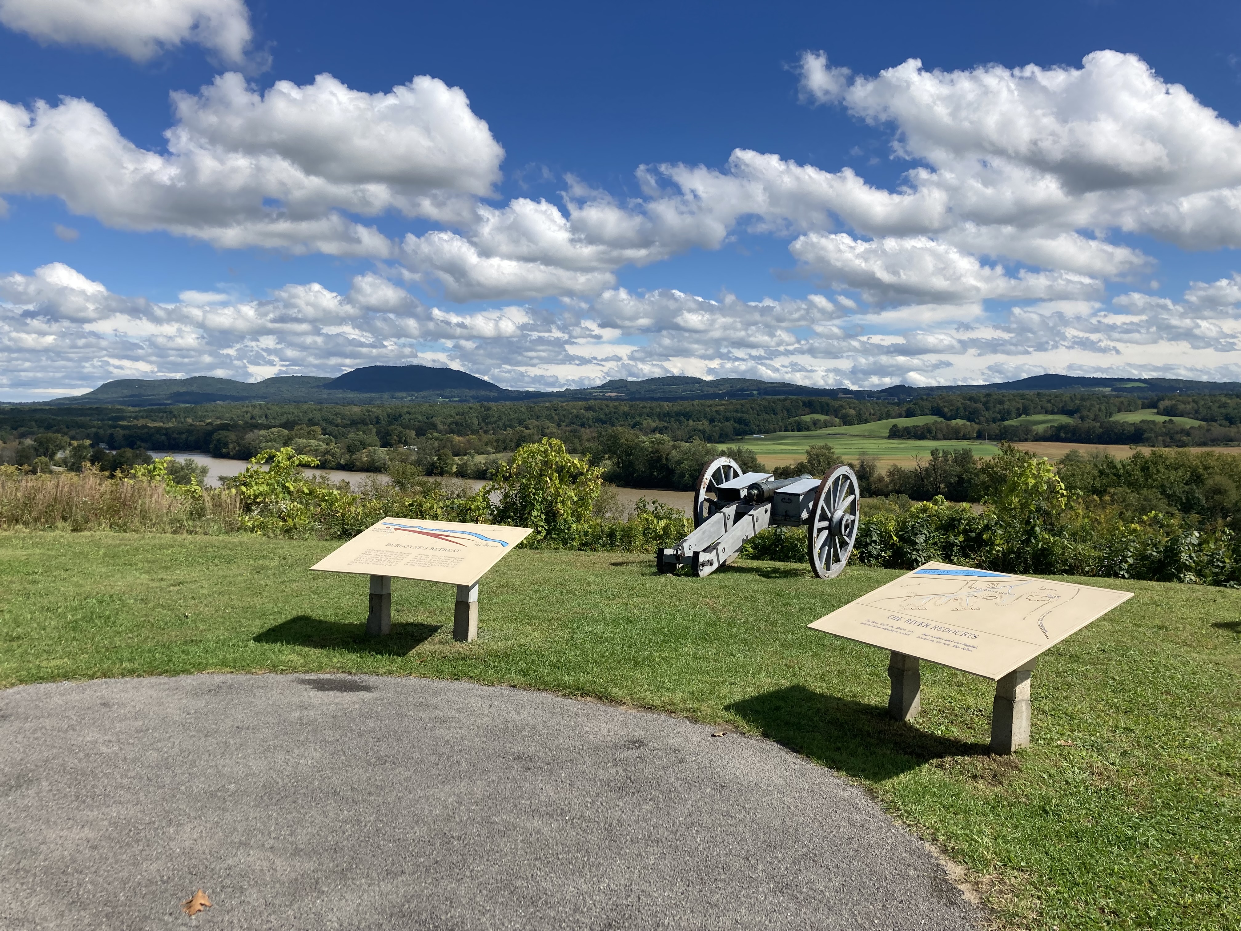 Panoramic view overlooking the river with green mountains in the background. A cannon is in the center flanked by two metal signs.
