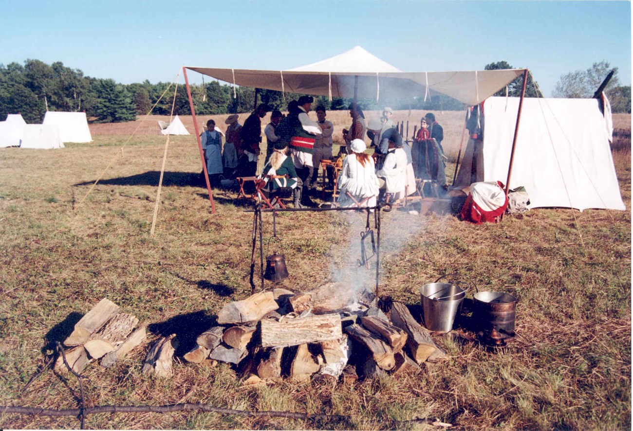 Members of the British camp gather under a large canvas tent. In the foreground is a campfire surrounded by a pile of logs