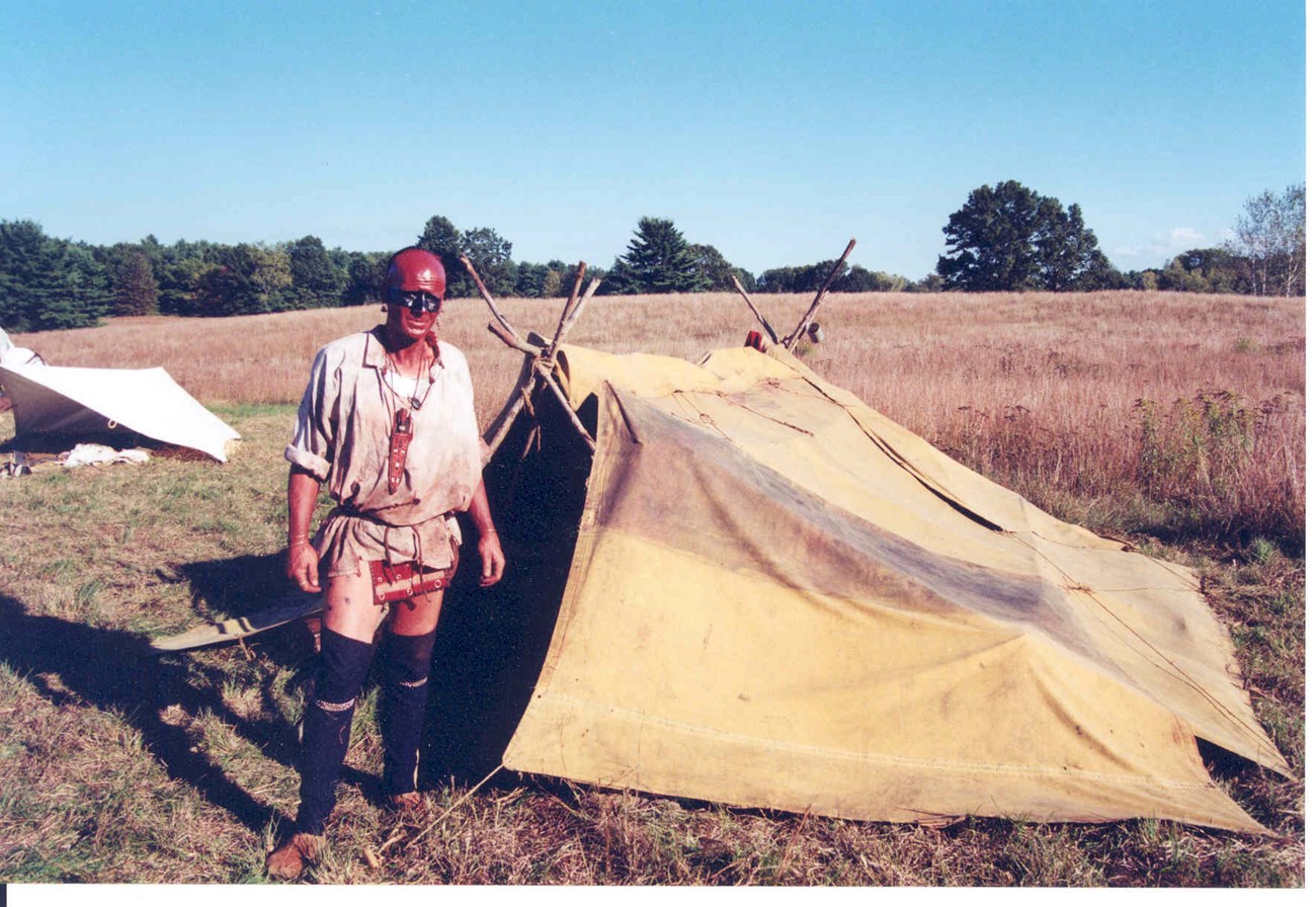 An Iroquois Native stands next to a brown tent in a field