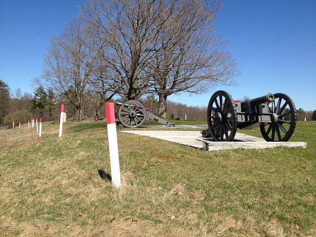 Red topped, white marker posts run in a line into the distance next to a cannon resting on a concrete pad.