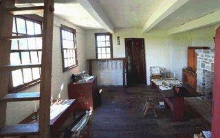 Interior view of the Neilson Farmhouse, a table and brick fireplace on the right, a ladder in the foreground on the left, and various tools throughout the room.
