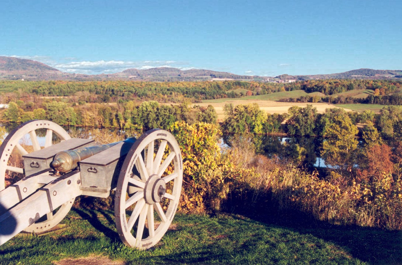 A cannon, with large wheels, rests on a grassy hill overlooking the Hudson River Valley with a clear blue sky overhead