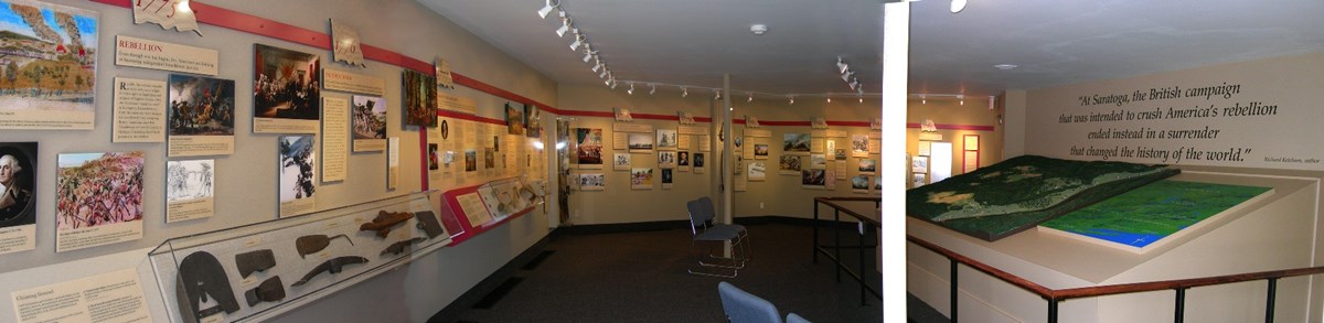 Panoramic of the Timeline Exhibit. The wall is covered with photos and text that runs along a timeline. artifacts, like weapons, are also among the exhibit