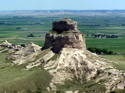 A large, cylindrical, sandstone formation rises above farmlands below.