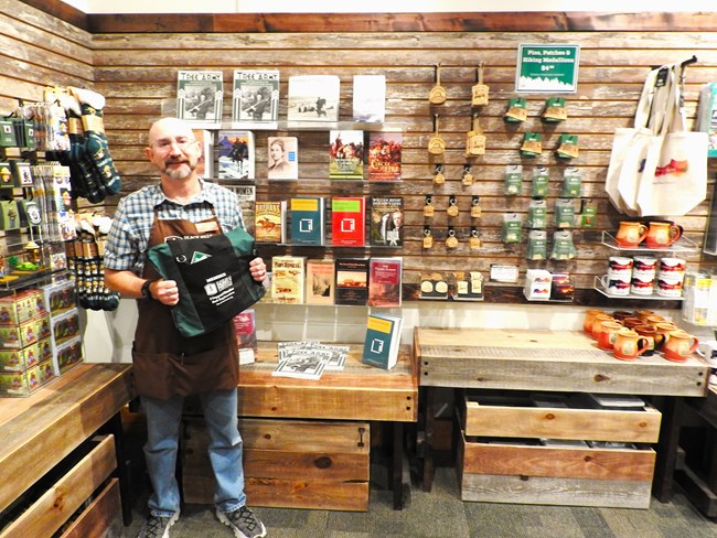 A man wearing an apron stands in front of a wall covered with books and other merchandise.
