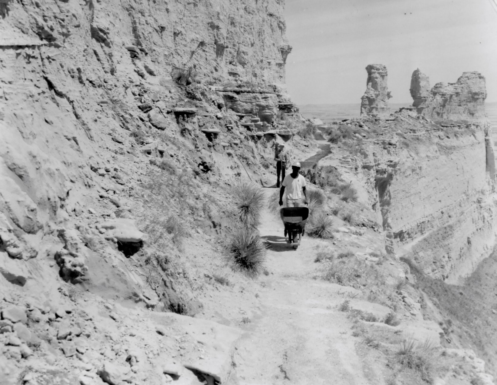 Scotts Bluff National Monument employees make repairs to the Saddle Rock Trail in 1971. Two men, one pushing a wheelbarrow, work on a trail that is perched on a ledge.
