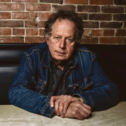 A man wearing a denim jacket is seated in front of a brick wall.