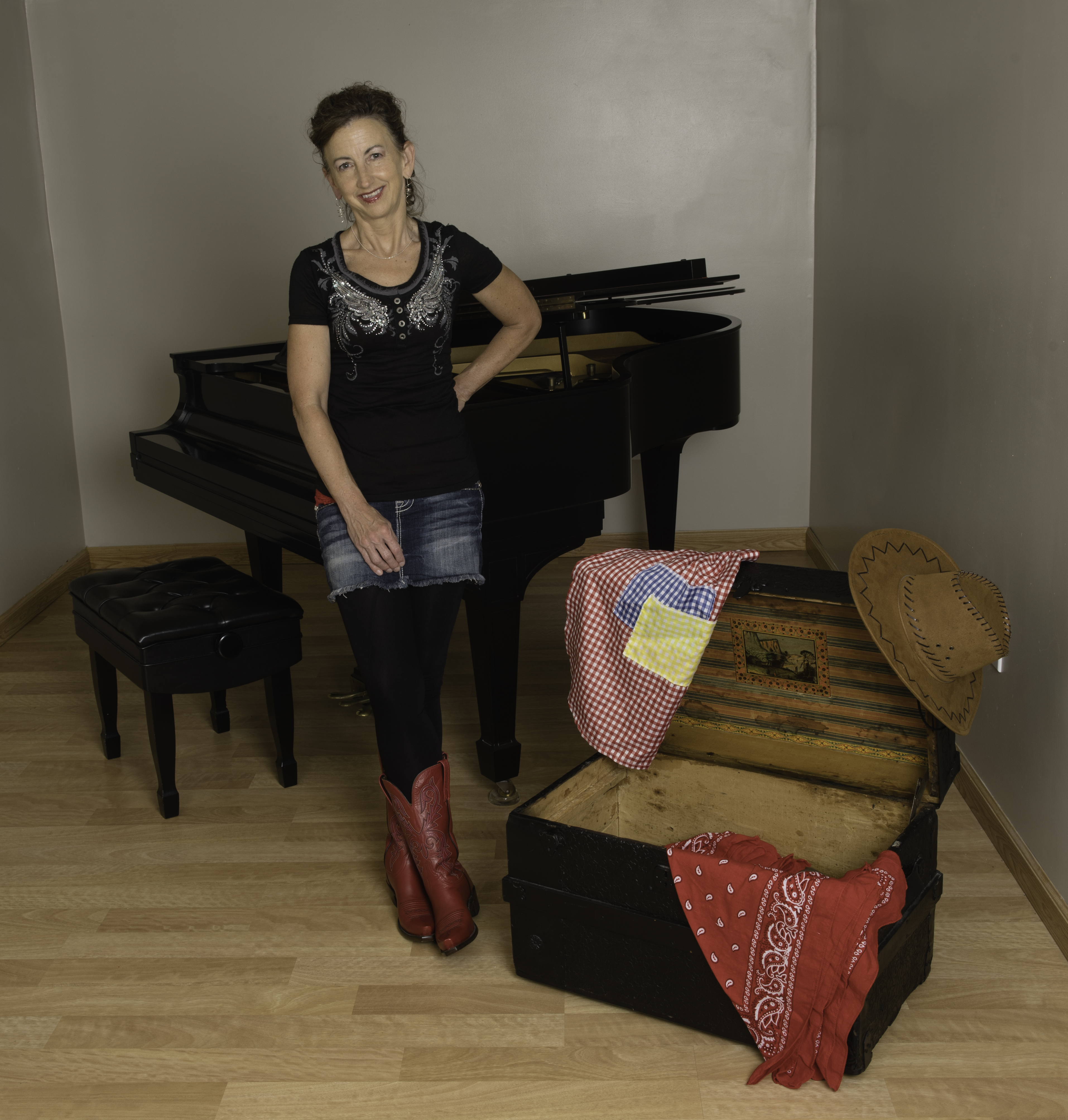 Donna Gunn will perform "Music on the Trail" online on Saturday, March 20, 2021 at 1 pm MDT. A woman leans against a piano with a trunk at her feet.