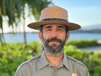 Jay Sturdevant A bearded man is seen from the chest up. He wears a park ranger hat. Water and vegetation are out of focus in the background.