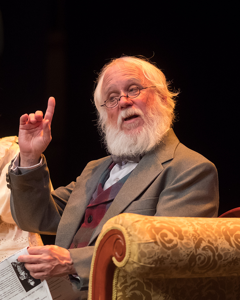 Brian Kokensparger acts in a stage production. A man holds a newspaper in one hand and points upwards with the other.