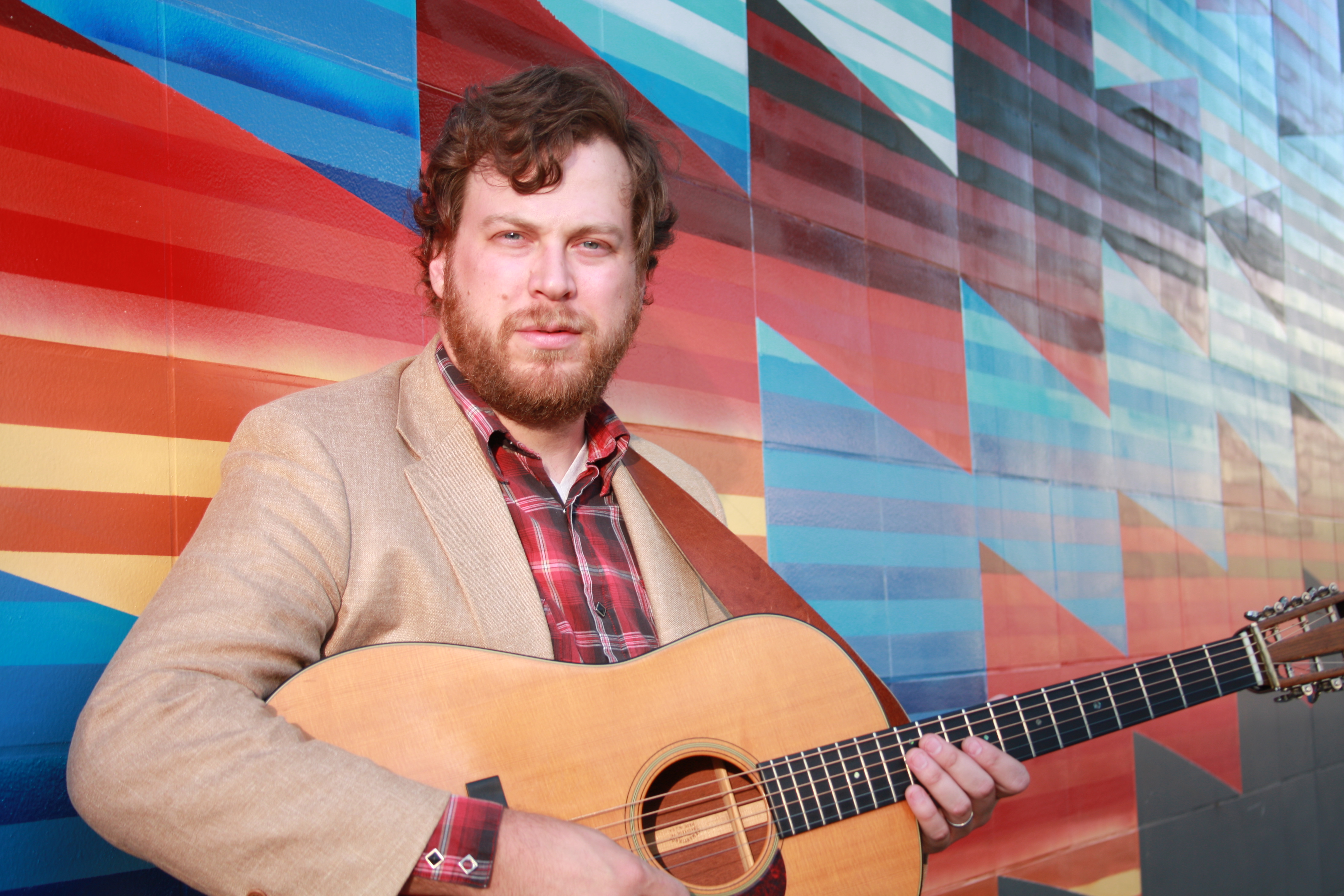A man holds a guitar in front of a colorful mural.